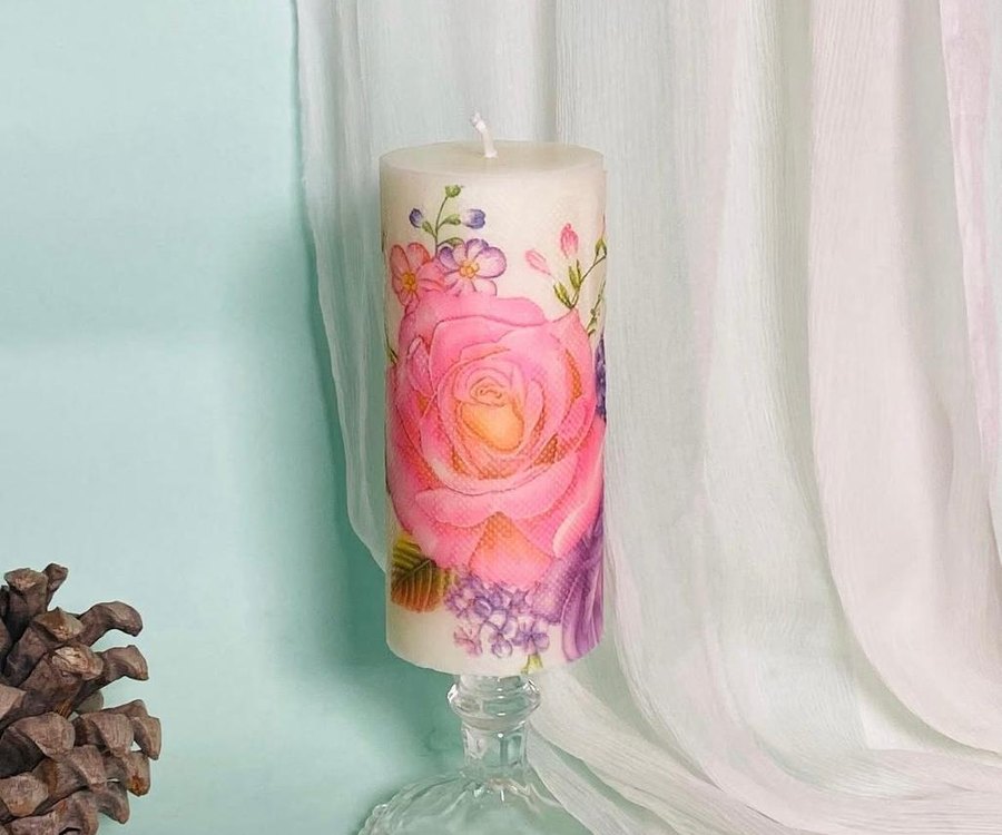 A pillar candle by Decor Studio by LA hand-painted with a pink rose and wildflowers, standing on a glass base beside a pine cone.